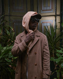 Person wearing a brown coat and pink hooded garment with black sunglasses, standing in front of a decorative door.